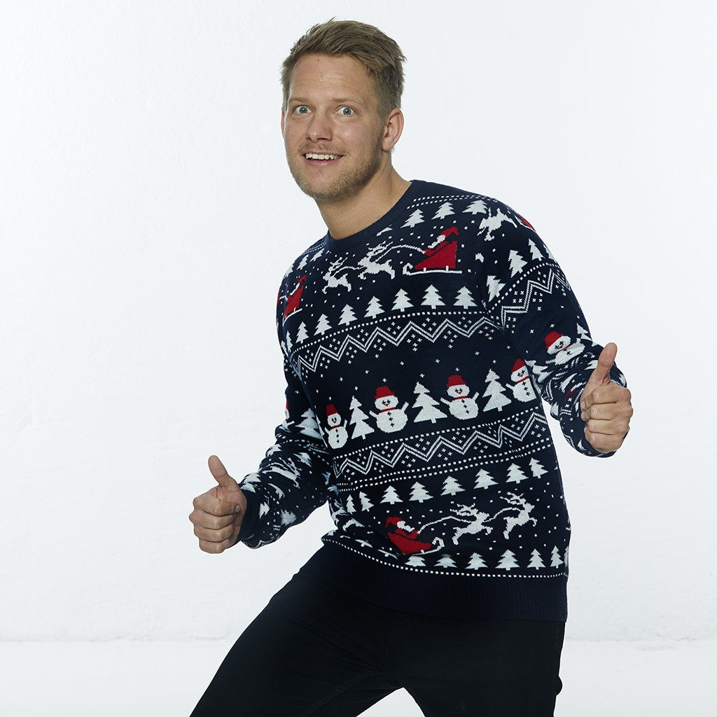 Un homme posant portant un pull de Noël avec des bonhommes de neige, des arbres de Noël et un motif de Noël.
